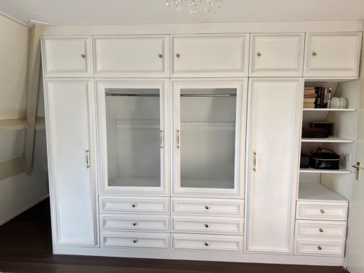 Witte wandkast met glazen deuren, lades en plankjes aan de zijkant.